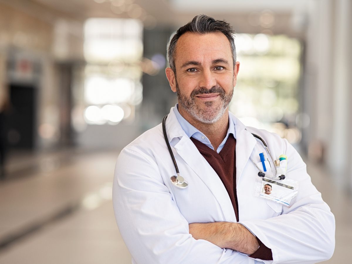 Male doctor standing in hallway with lab coat on and stethoscope around neck for article Finding a Doctor | Phoenix Physicians for newcomers moving to Phoenix.