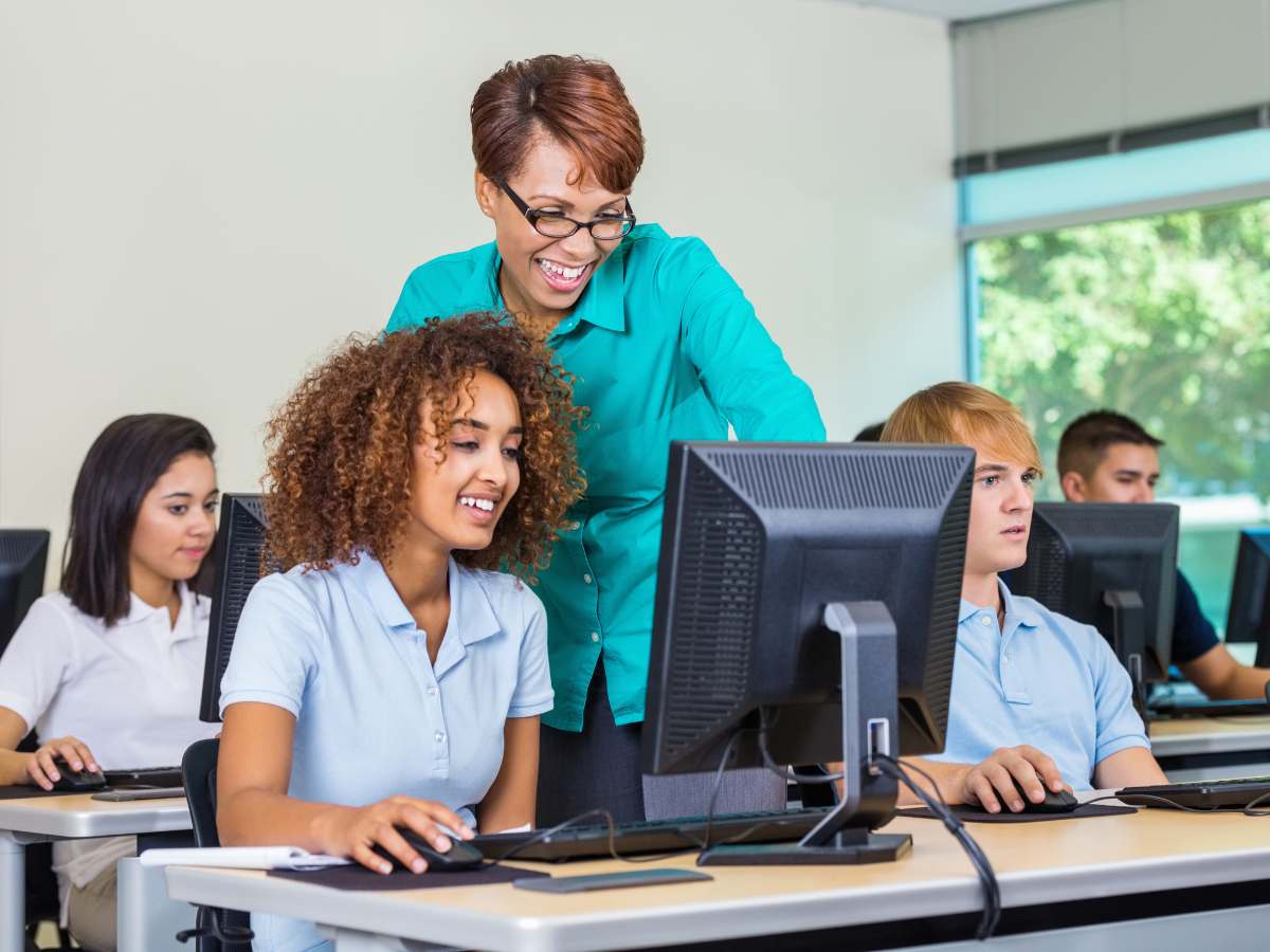 Classroom with private high sc;hool kids at desks with computers and teacher leaning over student to help for article 17 Best Private High Schools in Phoenix Metro for newcomers moving to Phoenix.