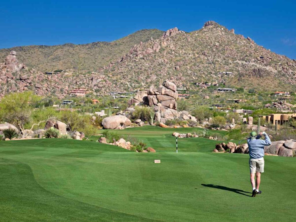 Man swinging golf club with desert mountains in background at golf course in Arizona for article Master Planned Communities in Phoenix for newcomers moving to Phoenix.