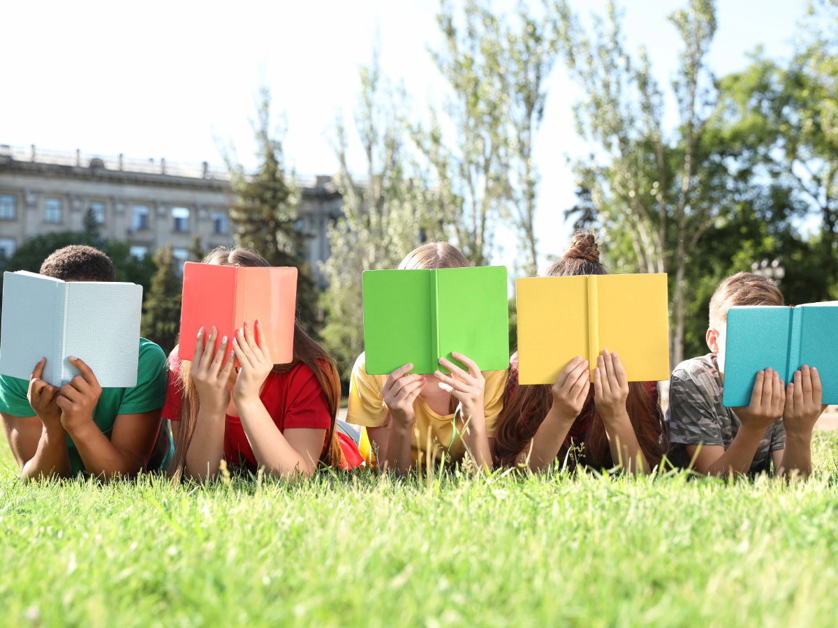 Children laying on the ground outside with books in front of their faces reading.