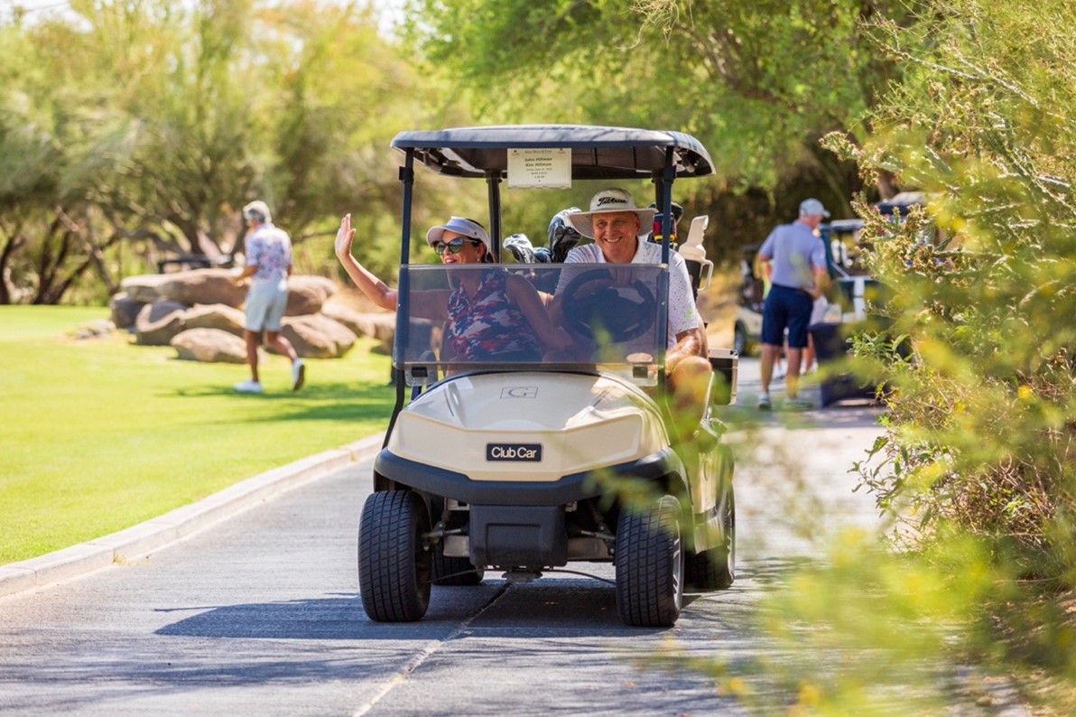 People smiling and waving in a golf cart driving on a path on the golf course.