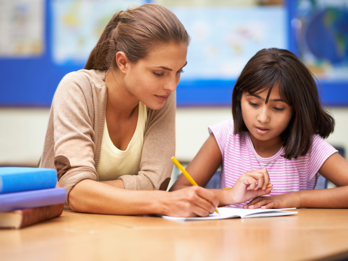 Female teacher sitting at a table writing in an open notebook while tutoring a young girl who is sitting next to her.