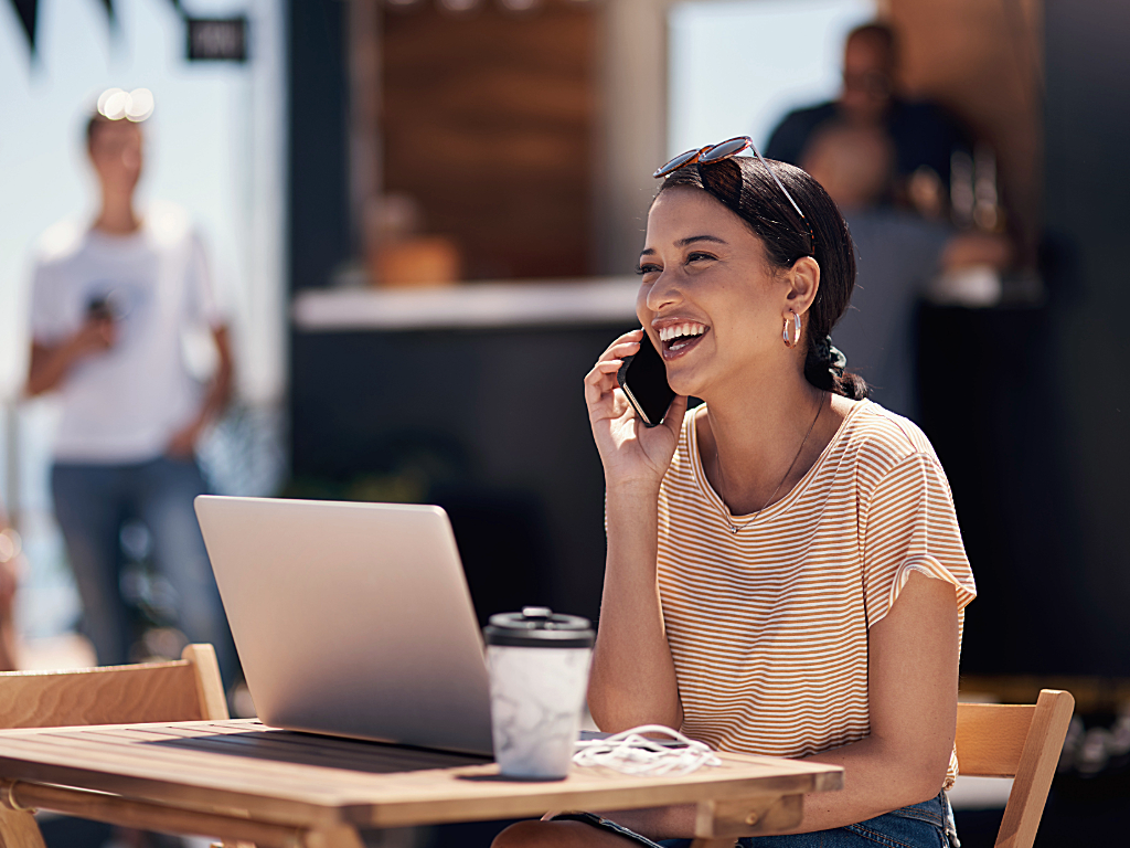 Woman on cell phone working working remote, with laptop and coffee on outdoor cafe table.