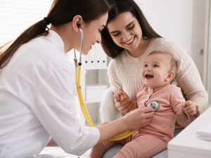 Pediatrician with stethoscope checking heartbeat of smiling baby girl while smiling mom holds her.