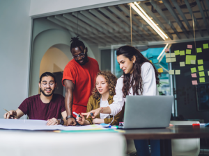 Group of 4 diverse people working on plans for startup company at desk with post it notes all over back wall.