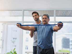 Physical therapist standing behind client helping to guide him as he raises both arms while holding resistance band.