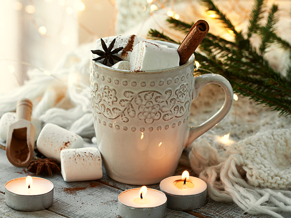 White coffee mug with coffee in it and topped with marshmellows and cinnamon sticks sitting on table with marshmellows, tea light candles, pine and cozy knit scarf around the cup for article The 12 Best Coffee Shops in Phoenix for Newcomers moving to Phoenix.