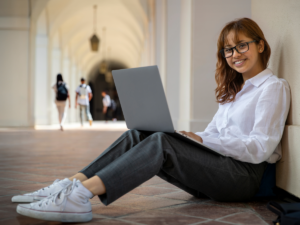 High school girl sitting on ground in school hallway with laptop open propped on her knees for article Top 4 Private High Schools in Phoenix for newcomers moving to Phoenix.