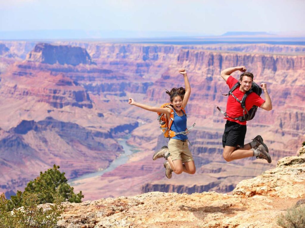 A male and a female with backpacks on jumping up in the air in front of the grand canyon for article Places to Hike Near Phoenix for newcomers moving to Phoenix.