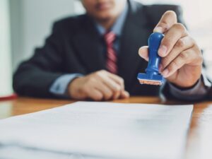 Blurred male in coat and tie sitting at table in background holding a rubber stamp over a piece of paper for article Navigating Professional Licensing in Phoenix for newcomers moving to Phoenix.