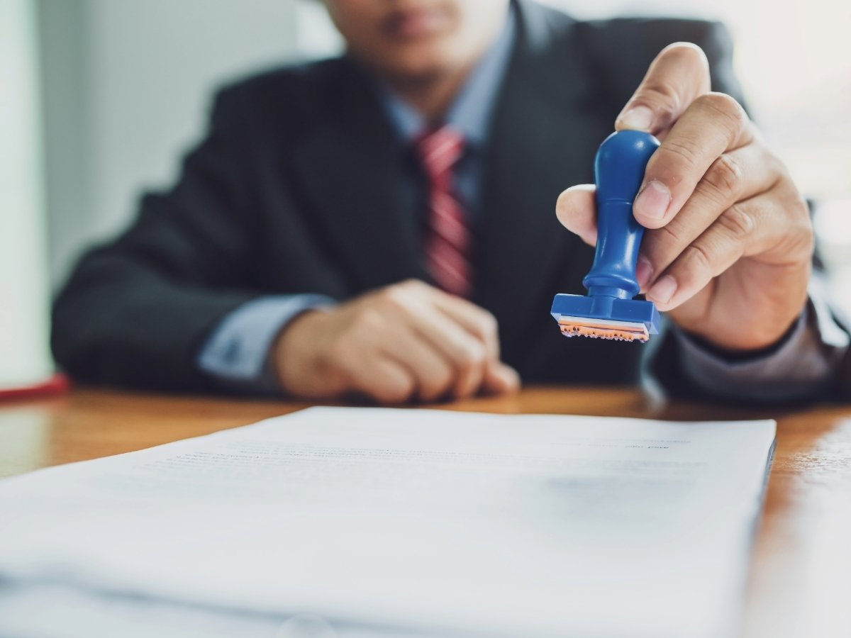 Blurred male in coat and tie sitting at table in background holding a rubber stamp over a piece of paper for article Navigating Professional Licensing in Phoenix for newcomers moving to Phoenix.