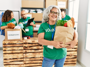 Senior woman wearing a green shirt that says volunteer - holding a sack of groceries and standing in front of a counter with boxes of donated items.